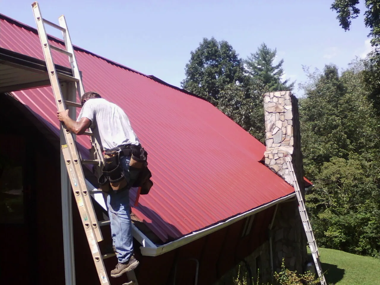 Roofer on a ladder beside a red metal roof and stone chimney — Soffit & Fascia Repair by Roofing Brothers in Joplin